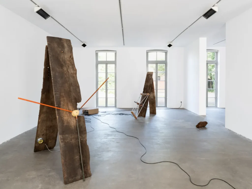 Zwei aufrechte, verwitterte Holzbalken mit orangefarbenen Stäben und Kabeln als Klanginstallation in einem weißen Galerieraum.