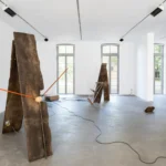 Zwei aufrechte, verwitterte Holzbalken mit orangefarbenen Stäben und Kabeln als Klanginstallation in einem weißen Galerieraum.