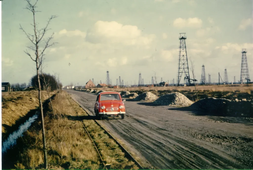 Roter Kleinwagen auf Schotterstraße inmitten zahlreicher Erdöl-Fördergerüste im Emsland der 1950er Jahre.