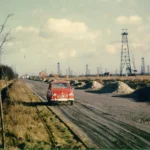 Roter Kleinwagen auf Schotterstraße inmitten zahlreicher Erdöl-Fördergerüste im Emsland der 1950er Jahre.