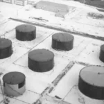 Luftaufnahme zeigt acht zylindrische Rohöltanks der Raffinerie Lingen-Holthausen auf ebenem Sandboden, im Hintergrund Fabrikgebäude.