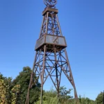 Metallener Bohrturm als Denkmal für den Ölboom in Dalum, mit historischen Symbolen, vor blauem Himmel.