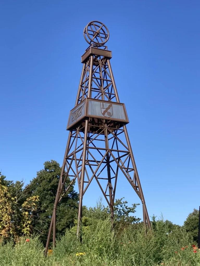 Metallener Bohrturm als Denkmal für den Ölboom in Dalum, mit historischen Symbolen, vor blauem Himmel.