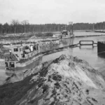 Schwarzweißfoto eines Schwimmbaggers beim Ausheben eines Kanals im Forstgebiet Lingen, mit Sandaufschüttungen im Vordergrund.