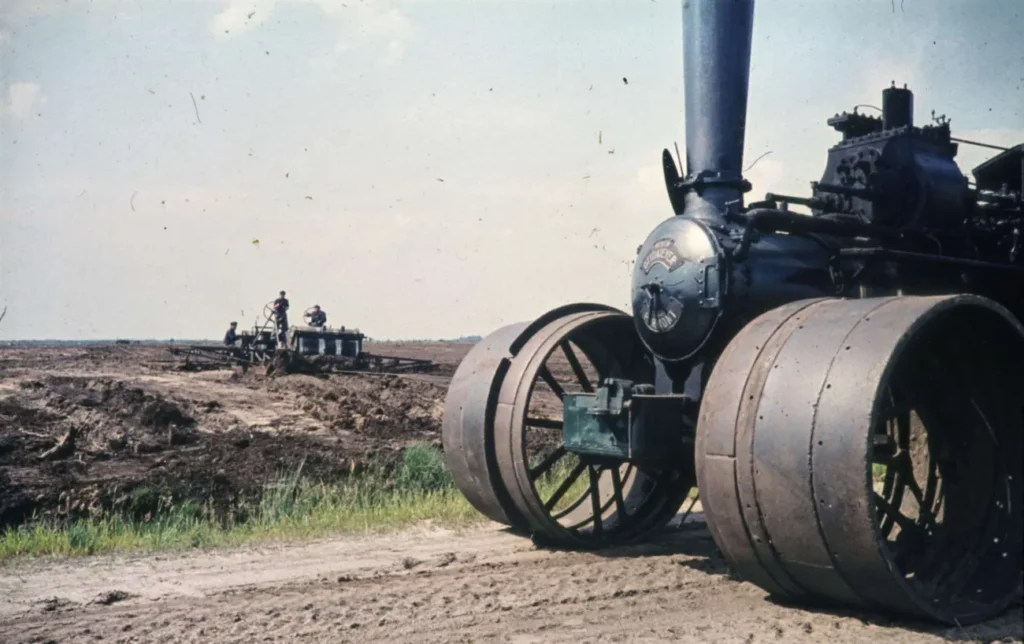 Eine historische Dampflokomobile steht auf abgetorftem Moorboden, im Hintergrund arbeiten Männer an einer Maschine.