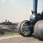 Eine historische Dampflokomobile steht auf abgetorftem Moorboden, im Hintergrund arbeiten Männer an einer Maschine.