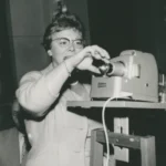 Eine Frau mit Brille bedient einen Diaprojektor auf einem Holzständer bei einem Vortrag, ca. 1950er Jahre.