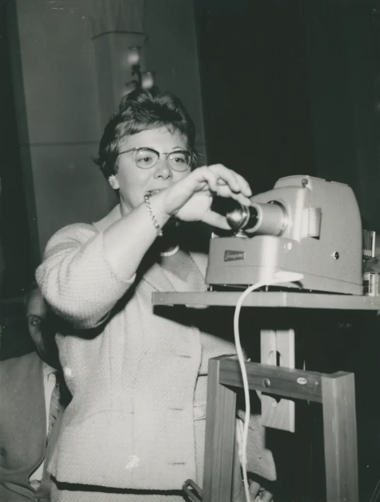 Eine Frau mit Brille bedient einen Diaprojektor auf einem Holzständer bei einem Vortrag, ca. 1950er Jahre.