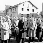 Männer in Mänteln stehen bei einer öffentlichen Veranstaltung auf einem Stadtplatz, umgeben von einer großen Menschenmenge.