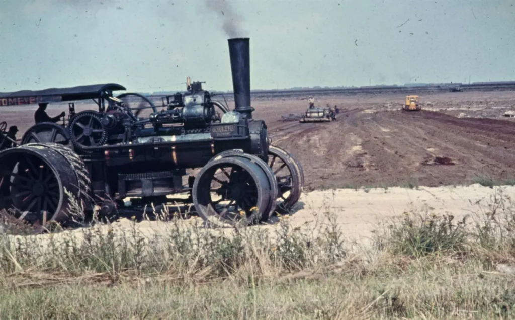 Eine historische Dampflokomobile steht auf einem abgetorften Moorgelände, im Hintergrund arbeiten weitere Maschinen auf dem Feld.