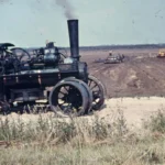Eine historische Dampflokomobile steht auf einem abgetorften Moorgelände, im Hintergrund arbeiten weitere Maschinen auf dem Feld.