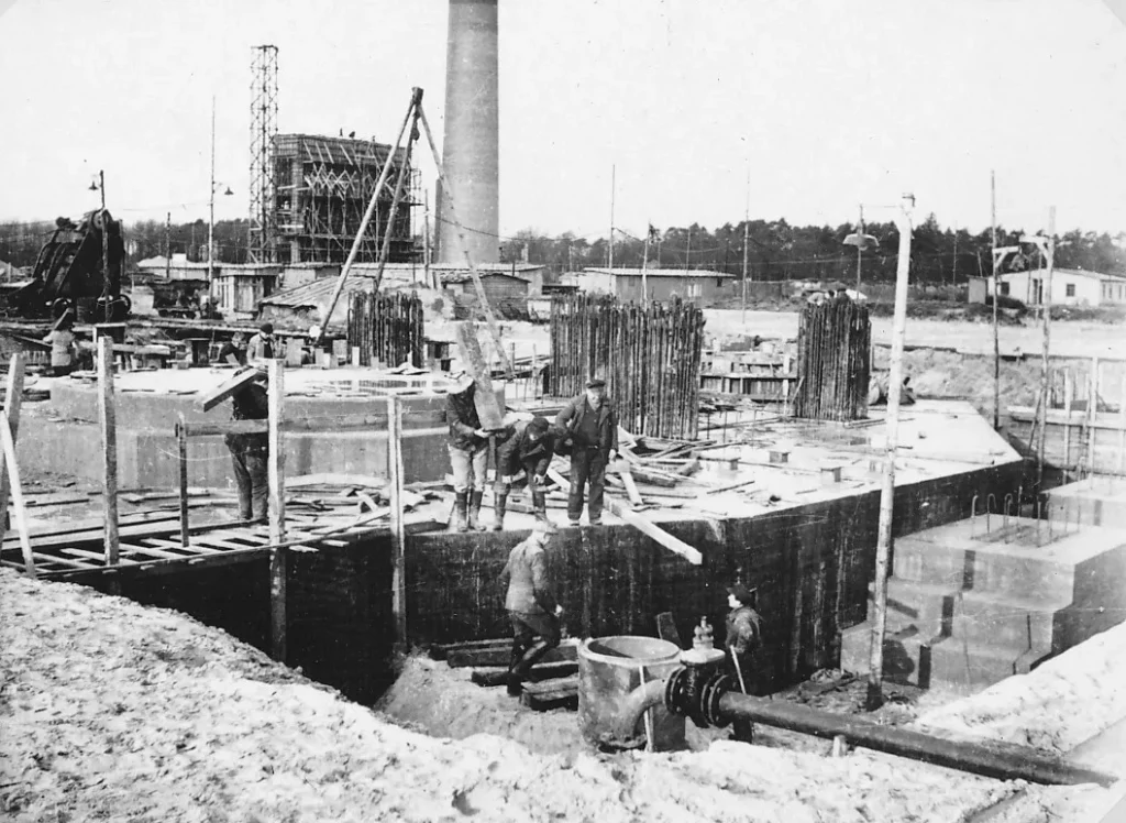 Arbeiter errichten Betonfundamente mit Stahlarmierungen auf einer Sandfläche, im Hintergrund ein Schornstein und Baugerüste.