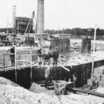 Arbeiter errichten Betonfundamente mit Stahlarmierungen auf einer Sandfläche, im Hintergrund ein Schornstein und Baugerüste.