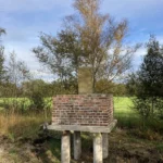 Historischer Grenzstein auf erhöhtem Backsteinpodest mit Holzpfählen, umgeben von Moorlandschaft und Herbstbäumen.