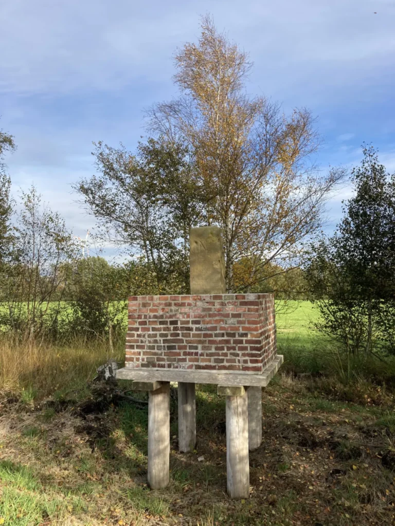 Historischer Grenzstein auf erhöhtem Backsteinpodest mit Holzpfählen, umgeben von Moorlandschaft und Herbstbäumen.