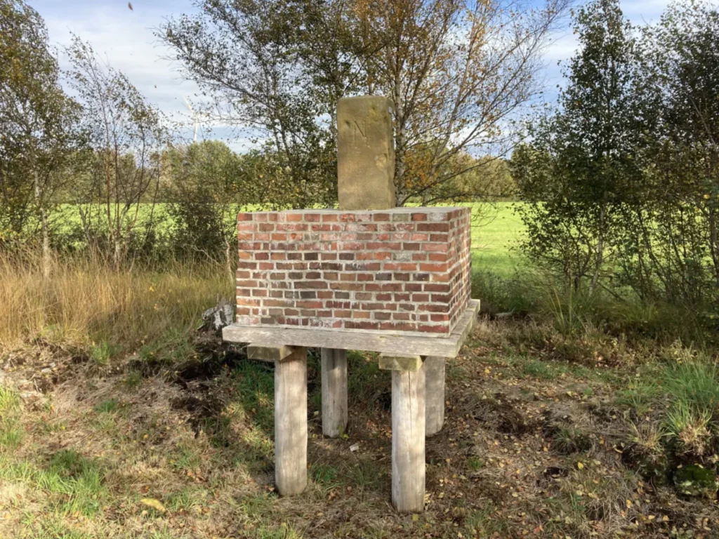 Ein alter Grenzstein auf einem Backsteinpodest, das auf Holzpfählen über dem Moorbodenniveau steht.