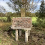 Ein alter Grenzstein auf einem Backsteinpodest, das auf Holzpfählen über dem Moorbodenniveau steht.