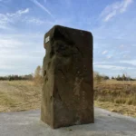 Ein alter, verwitterter rechteckiger Grenzstein aus dem 18. Jahrhundert steht auf einem Betonsockel in flacher Moorlandschaft.