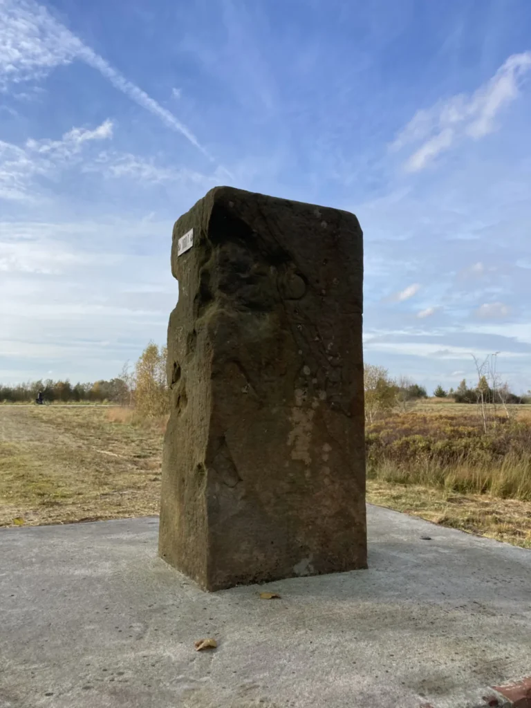 Ein alter, verwitterter rechteckiger Grenzstein aus dem 18. Jahrhundert steht auf einem Betonsockel in flacher Moorlandschaft.