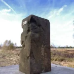 Alter Grenzstein aus dem 18. Jahrhundert auf einem Betonsockel in flacher Moorlandschaft unter blauem Himmel.
