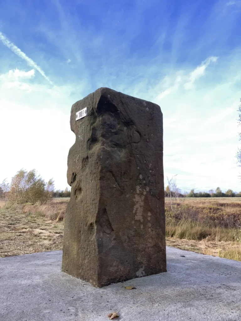 Alter Grenzstein aus dem 18. Jahrhundert auf einem Betonsockel in flacher Moorlandschaft unter blauem Himmel.