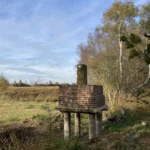 Ein alter Grenzstein auf einem Backsteinpodest mit Holzbeinen steht am Rand eines Moorgebiets mit Birken.