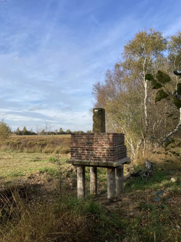 Ein alter Grenzstein auf einem Backsteinpodest mit Holzbeinen steht am Rand eines Moorgebiets mit Birken.