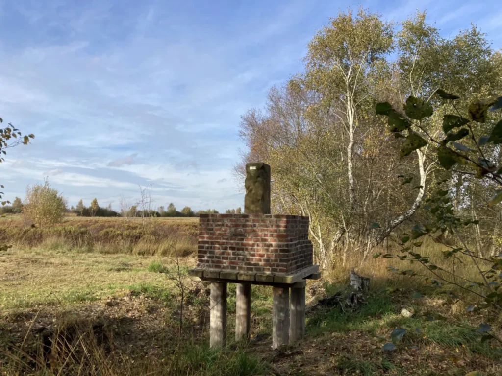 Ein alter Grenzstein auf einem erhöhten Ziegelsockel mit Holzpfählen steht in einer moorigen Landschaft mit Birken.