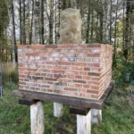 Ein alter Grenzstein aus Sandstein auf einem erhöhten Ziegelsockel, der auf Holzpfählen steht, um das Moorbodenniveau auszugleichen.