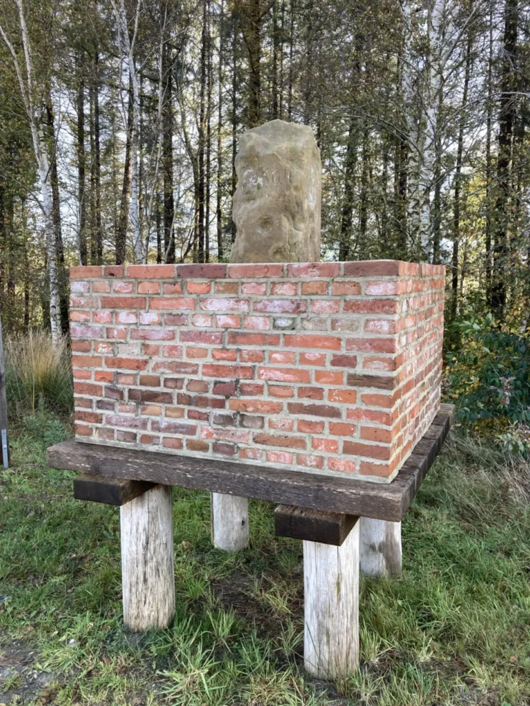 Ein alter Grenzstein aus Sandstein auf einem erhöhten Ziegelsockel, der auf Holzpfählen steht, um das Moorbodenniveau auszugleichen.