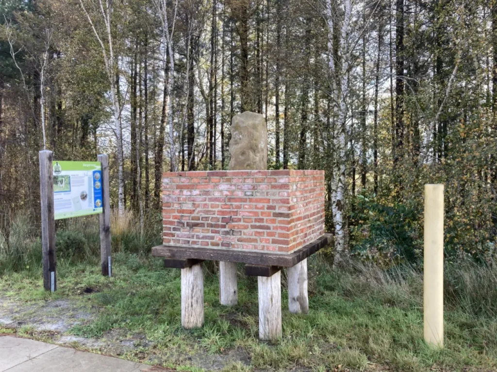 Ein alter Grenzstein auf einem erhöhten Backsteinpodest mit Holzstützen, umgeben von Wald und einer Informationstafel.