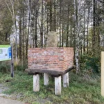 Ein alter Grenzstein auf einem erhöhten Backsteinpodest mit Holzstützen, umgeben von Wald und einer Informationstafel.