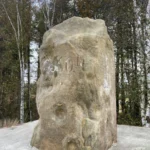 Ein verwitterter Sandstein-Grenzpfeiler auf einem Betonsockel, umgeben von Birken und Nadelbäumen, mit kaum lesbarer Inschrift.