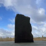 Dunkler Grenzstein auf einem Steinsockel vor weitem Moorlandschaftshorizont und bewölktem Himmel.