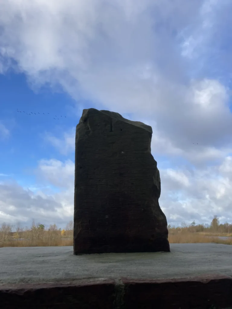Dunkler Grenzstein auf einem Steinsockel vor weitem Moorlandschaftshorizont und bewölktem Himmel.