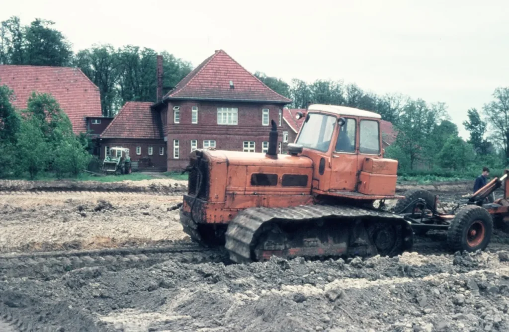 Ein orangefarbener Raupenbagger arbeitet auf schlammigem Feld bei der Flurbereinigung, im Hintergrund ein Backsteingebäude.