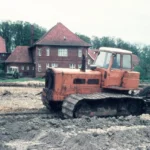 Ein orangefarbener Raupenbagger arbeitet auf schlammigem Feld bei der Flurbereinigung, im Hintergrund ein Backsteingebäude.