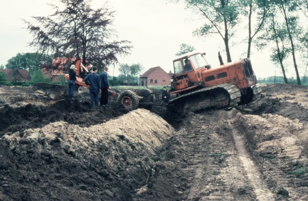 Eine orange Planierraupe und Arbeiter bei Erdarbeiten zur Flurbereinigung im schlammigen Gelände, 1960er Jahre.