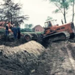Eine orange Planierraupe und Arbeiter bei Erdarbeiten zur Flurbereinigung im schlammigen Gelände, 1960er Jahre.