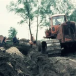 Ein orangefarbener Bulldozer planiert schlammiges Gelände, begleitet von mehreren Arbeitern bei Flurbereinigungsarbeiten im Emsland.