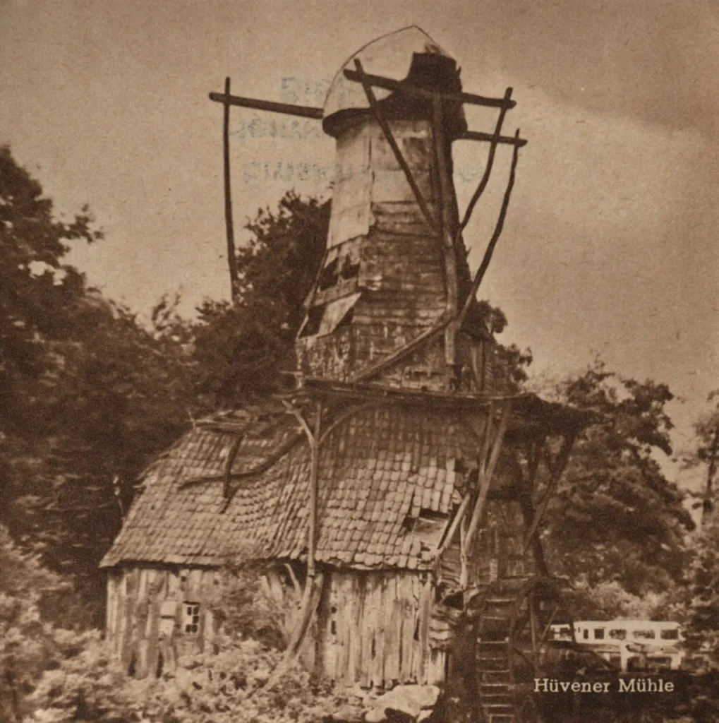 Historische Aufnahme der verfallenen Hüvener Mühle, ein altes Holzgebäude mit baufälligem Windmühlenaufsatz inmitten von Bäumen.