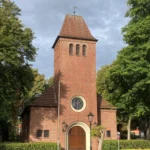 Backsteinkirche mit quadratischem Turm, Rundfenster und Holzportal, umgeben von Hecken und Bäumen in Hasselbrock.
