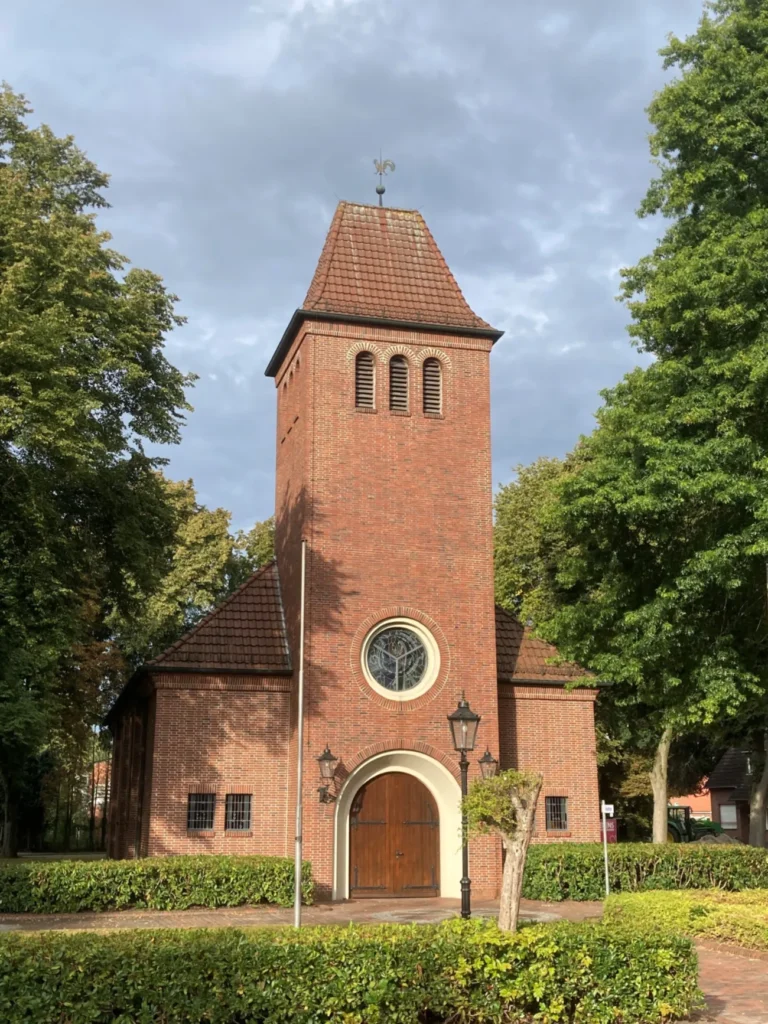 Backsteinkirche mit quadratischem Turm, Rundfenster und Holzportal, umgeben von Hecken und Bäumen in Hasselbrock.