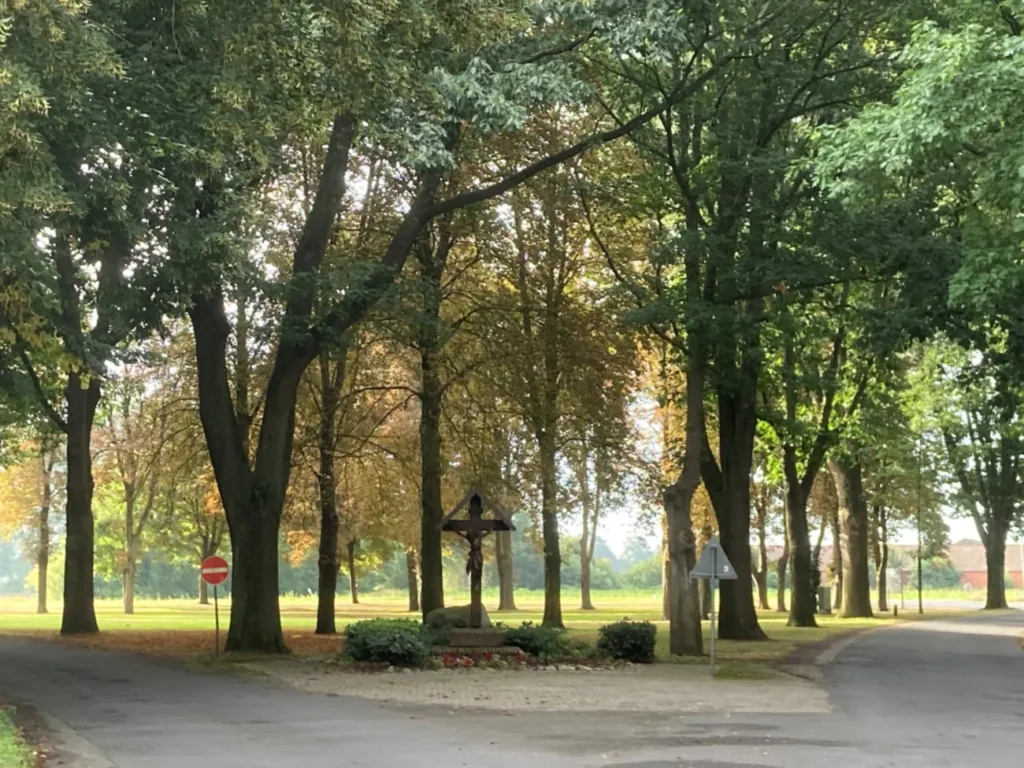 Holzkreuz mit Christusfigur an einer Dorfstraße, umgeben von alten Bäumen und grünen Wiesen.