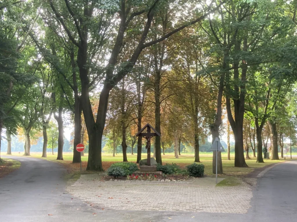 Hölzernes Wegkreuz mit Korpus auf gepflastertem Platz, umgeben von Bäumen einer Alleen-Kreuzung.