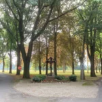 Hölzernes Wegkreuz mit Korpus auf gepflastertem Platz, umgeben von Bäumen einer Alleen-Kreuzung.