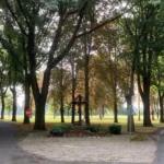 Hölzernes Wegkreuz mit Korpus auf gemauertem Sockel, umgeben von Büschen und roten Kerzen, in einer Baumallee.