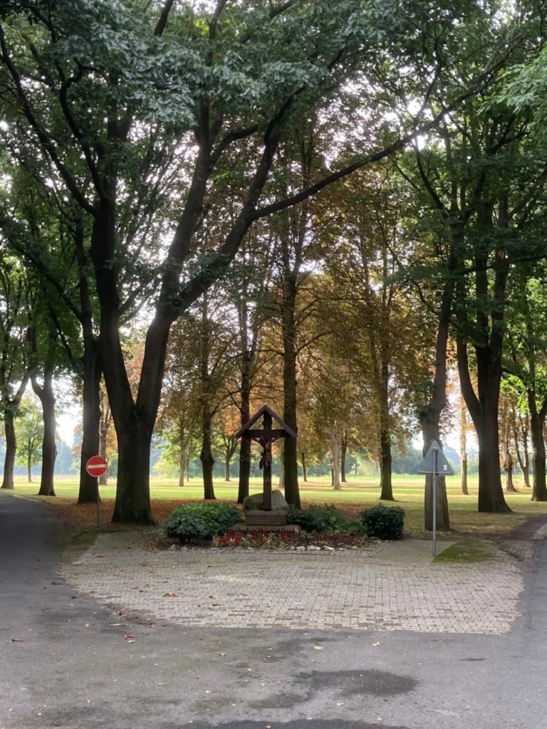 Hölzernes Wegkreuz mit Korpus auf gemauertem Sockel, umgeben von Büschen und roten Kerzen, in einer Baumallee.