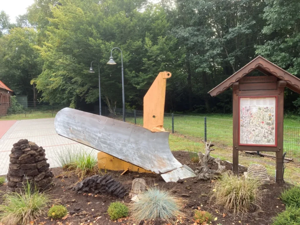 Ein gelb-silberner Schneepflug steht als Denkmal in einem bepflanzten Beet neben einer Informationstafel im Dorf Hasselbrock.