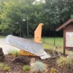 Ein gelb-silberner Schneepflug steht als Denkmal in einem bepflanzten Beet neben einer Informationstafel im Dorf Hasselbrock.